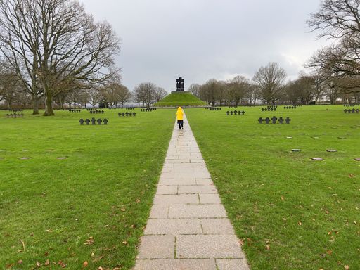 Child in a yellow jacket walks along a path through a cemetery with a monument in the distance, surrounded by bare trees and green grass.