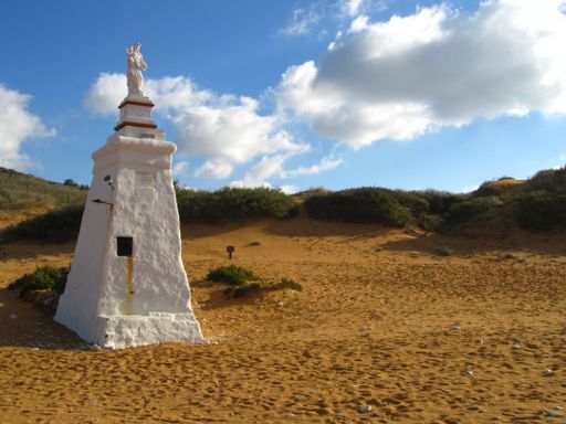 A statue of the Virgin Mary on a big white pedestal in the beautiful Ramla l-Hamra Bay in the island of Gozo, with its orange sand and cloudy sky. Mal