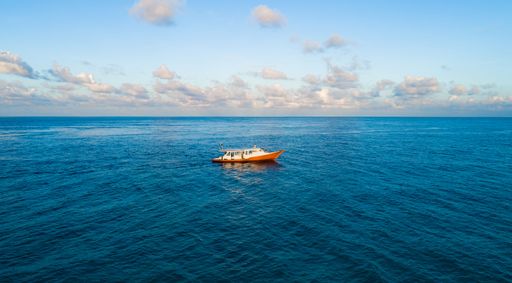 A lone boat sails on the vast blue ocean under a bright sky with scattered clouds, capturing the serenity of open waters.