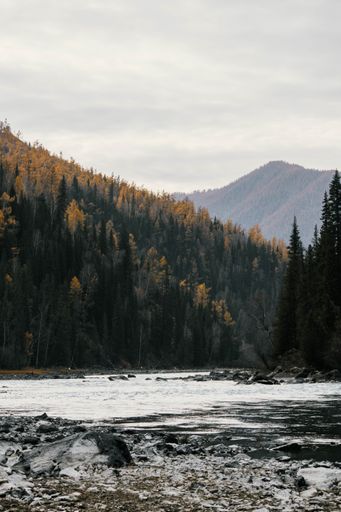 Scenic view of a flowing river with rocky banks and autumn forested hills under a cloudy sky.