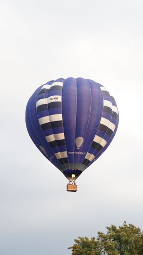 A vibrant blue hot air balloon with white stripes floating above trees under a clear sky.