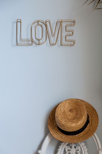 A vertical shot of a straw hat hanging on the white chair back leaned on the wall and a "Love" sign hanging on it