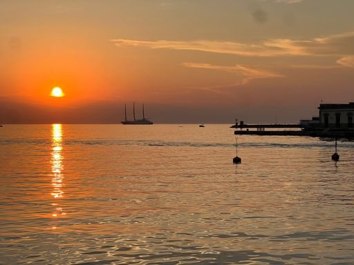 Serene sunset over a calm sea with a silhouetted sailboat and distant pier, creating a tranquil coastal scene.
