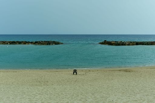 A serene beach scene with a lone chair on the sandy shore, calm turquoise waters, and a rocky breakwater under a clear sky.
