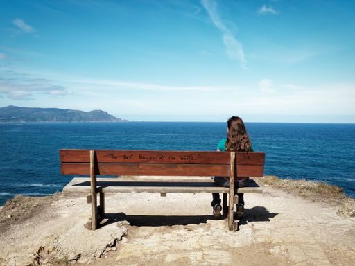 Woman sitting on a bench overlooking the ocean with a view of distant mountains under a clear blue sky.