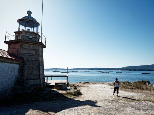 A woman stands near a lighthouse overlooking a calm sea under a clear blue sky, capturing the serene coastal landscape.