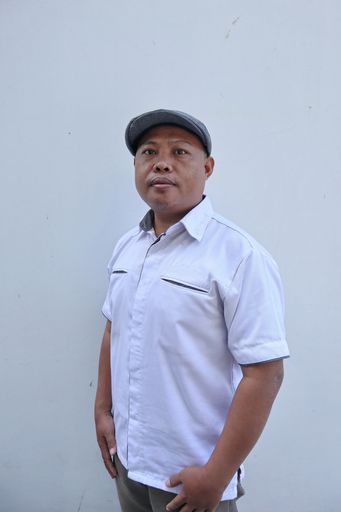 A vertical shot of a middle-aged Indonesian man standing in front of a white wall