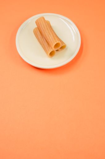 A vertical shot of rigatoni on a little white plate on an orange surface with free space for text