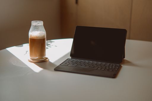 A closeup of a black tablet with a keyboard attached to it next to a bottle of iced coffee on a desk
