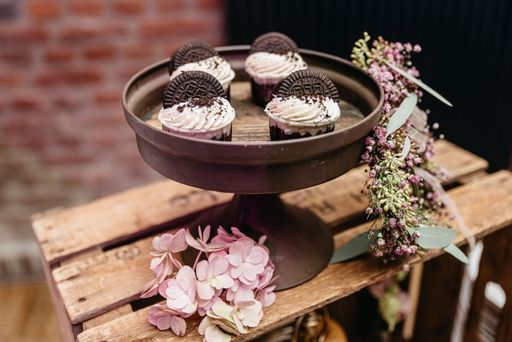 The cupcakes topped with chocolate cookies on a rustic stand, adorned with pink and purple flowers, set against a blurred brick background.