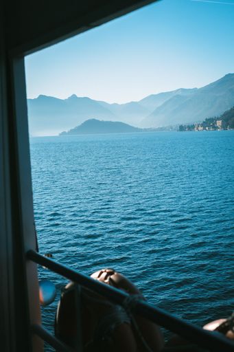 View from a boat window of serene Lake Como with distant mountains under a clear blue sky.
