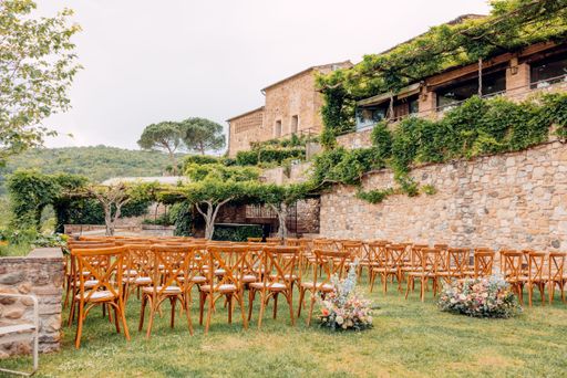 Charming outdoor wedding venue with wooden chairs and floral arrangements set against a rustic stone building and lush greenery.