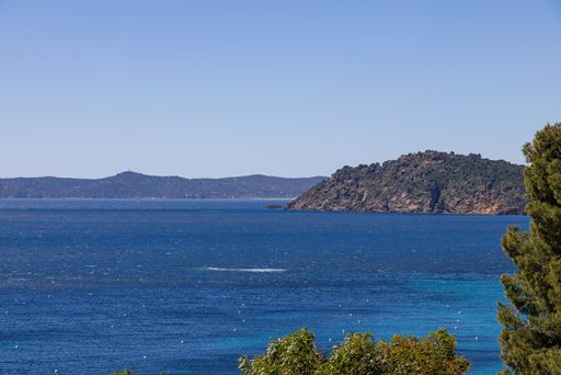 Scenic view of a clear blue sea with a rocky island and distant hills under a bright sky, framed by lush green foliage.