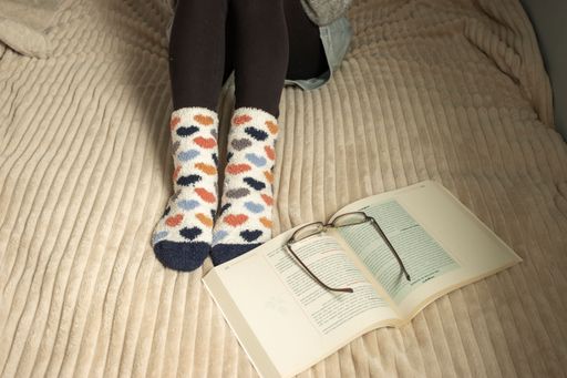 A woman stops reading taking off a pare of glasses. She uses fluffy a blanket and socks to warm up.