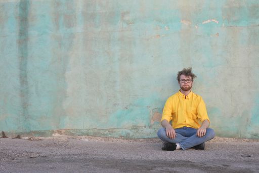 A young Hungarian ginger man with a beard and sunglasses sitting on a walkway against a wall outdoors