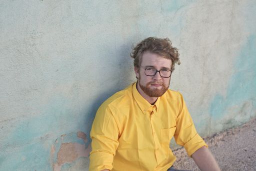 An adult Hungarian ginger man with glasses sitting on the ground and leaning on a wall outdoors