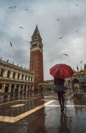A shot of beautiful architecture of the Tower of San Marco and a female walking with a red umbrella in Venice, Italy