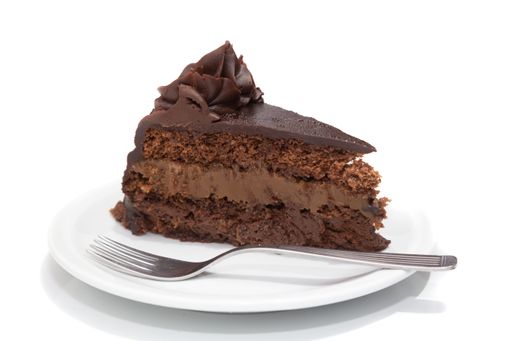 A slice of rich chocolate cake with creamy layers on a white plate, accompanied by a fork, set against a white background.