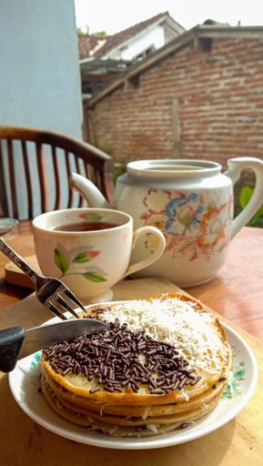 A closeup shot of a martabak with a chocolate topping and grated cheese and a cup of black tea with teapot