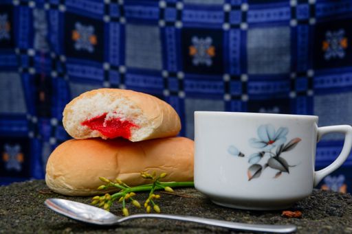 A close up of bread with strawberry jam with a cup of tea and a spoon with blue decorated background