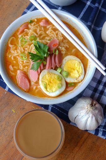 A vertical shot of a bowl full of noodle soup with toppings such as sausages, eggs, and vegetables