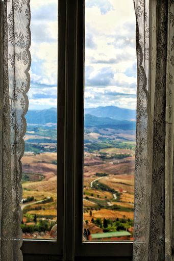 Scenic view of rolling hills through a window with lace curtains, capturing the beauty of the countryside under a cloudy sky.