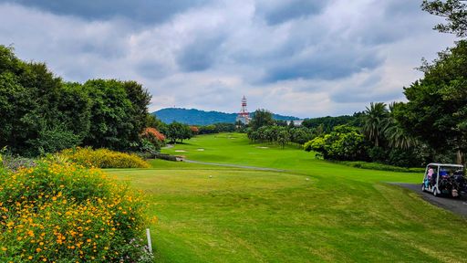 Scenic view of a lush green golf course with vibrant flowers and a cloudy sky, featuring a distant tower and golf cart.