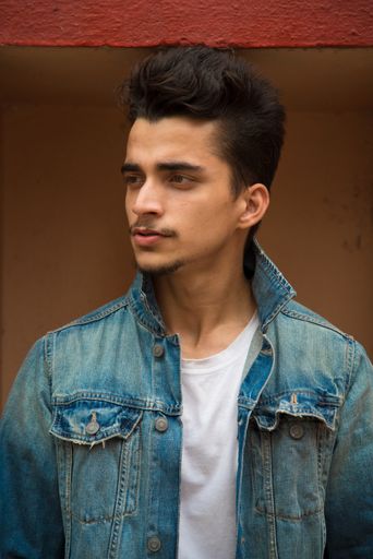 An attractive South Asian male posing in front of a brown wall wearing a jean jacket