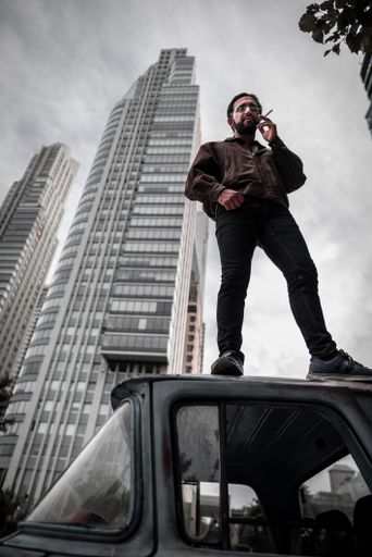 A Hispanic guy holding a cigarette and standing on a car with a skyscraper on the background