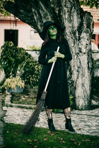 A vertical shot of a beautiful young witch with a broom and a Halloween costume in the garden