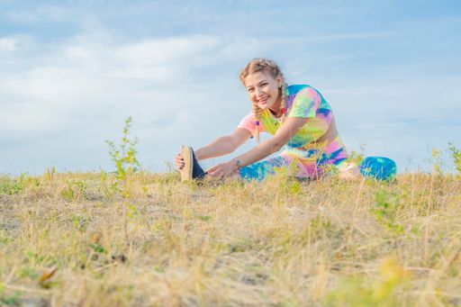 Young beautiful girl in bright tracksuit sits on green grass against blue sky and performs physical exercises to stretch muscles. Outdoor sports.
