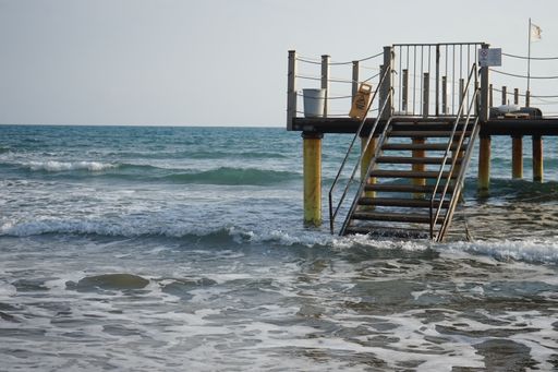 A close-up shot of a bridge with stairs over the sea with foamy waves rolling and crashing on a sandy beach