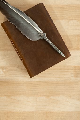 A decorated feather quill pen on an old handmade leather notebook isolated on a wooden desk mockup