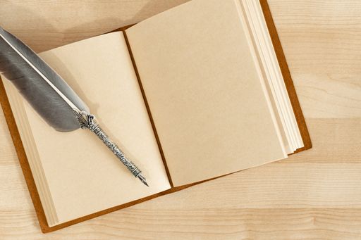 A top view of a feather quill pen with an old handmade leather notebook on a wooden desk