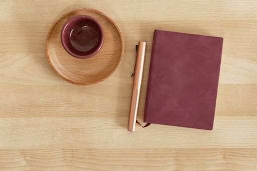 A top view of a burgundy notebook and pen, next to cup and wooden dish, on wooden desk