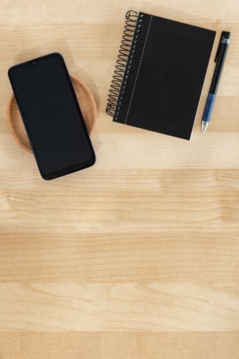 A vertical top view of a black smartphone, notebook, and pen on a wooden background, office banner style