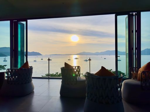 A view of a serene ocean sunset from a cozy indoor lounge with open doors, boats in the distance.