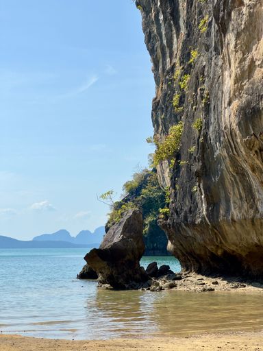 A view of a rocky cliff by a beach with clear blue waters and distant islands under a bright sky.