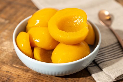Close up of a bowl full of canned peach halves on rustic wooden table with napkin and spoon