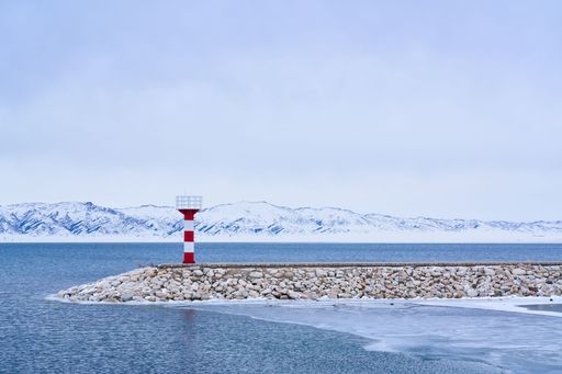 A serene winter scene of a red and white lighthouse on a rocky pier with snowy mountains and a calm sea under a cloudy sky.