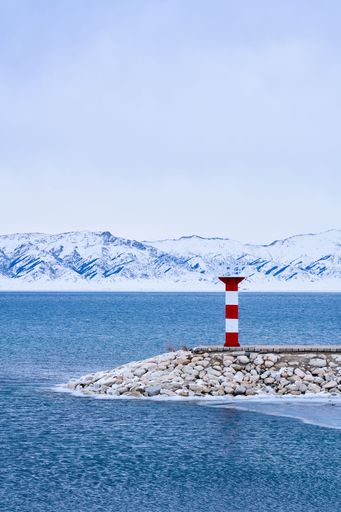 Scenic view of a red and white lighthouse on a rocky pier with icy mountains and a calm blue sea under a cloudy sky.
