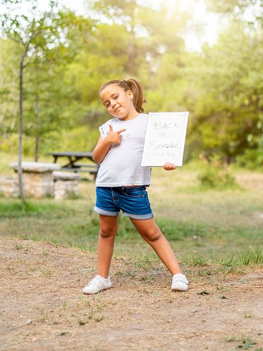 A smiling Caucasian girl in a park pointing to a blackboard reading "back to school".