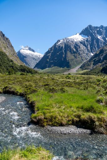 Scenic view of a mountain range with snow-capped peaks and a flowing stream in a lush green valley under a clear blue sky.