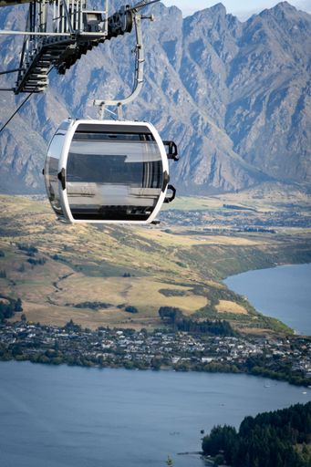 Scenic view of a gondola lift with majestic mountain backdrop in Queenstown, New Zealand, overlooking a serene lake and town.