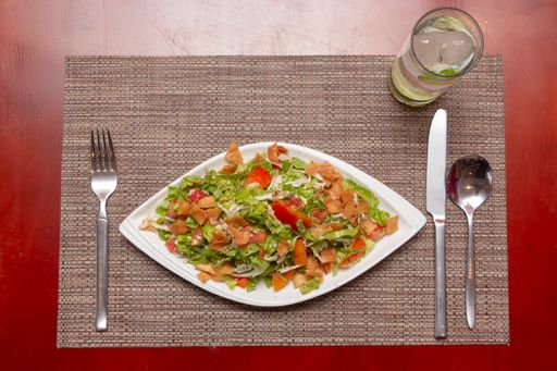 A top view shot of a green salad in a white dish with a glass of iced water on a light brown table