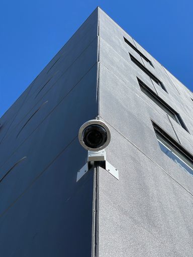 A closeup of a surveillance camera mounted on the edge of a building against a blue sky