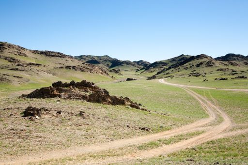 A scenic view of a winding dirt road through a grassy valley with rocky hills under a clear blue sky.
