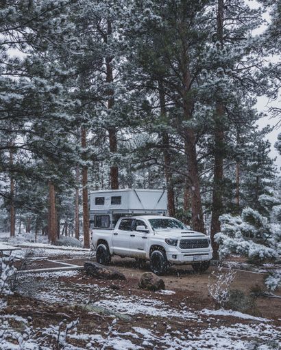 A white truck with a camper parked in a snowy forest in Estes Park, United States