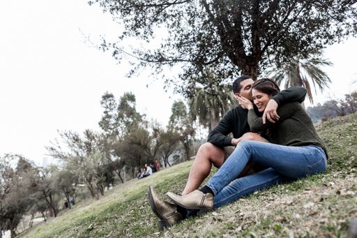 The Hispanic male kissing his girlfriend's head while sitting together on grassy field of a park