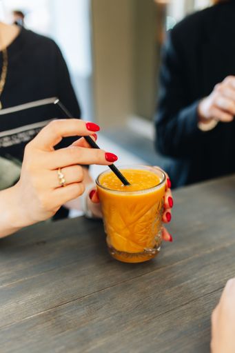 Close-up of a woman's hand with red nails holding a glass of orange juice at a cafe table.